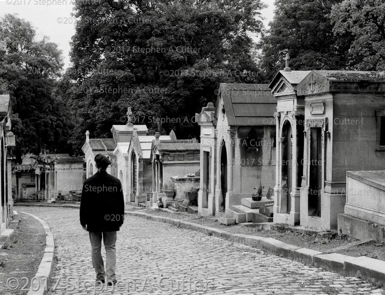 Black and White photograph of man in black beret walking away from camera, past a row of tombs in Pere Lachaise Cemetery Paris France
